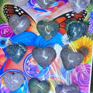 May include: A collection of 11 heart-shaped purple, green, and grey agate stones. The stones are arranged on a colorful background with a butterfly and flowers.