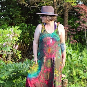 Może przedstawiać: Osoba w tie-dye kombinezonie w zielono-czerwono-żółtej kolorystyce. Kombinezon ma szerokie nogawki i luźny krój. Osoba nosi brązowy kapelusz i srebrny naszyjnik.