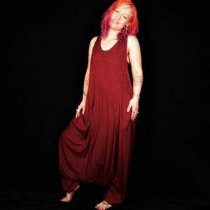 May include: A woman wearing a maroon jumpsuit with a scoop neck and sleeveless design. She has pink and red hair and is standing against a black background.