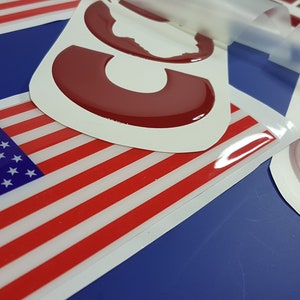 Op de afbeelding: Close-up van een rood, wit en blauwe Amerikaanse vlag sticker met een transparante beschermlaag. De sticker bevindt zich op een blauwe ondergrond.
