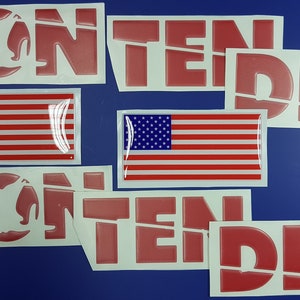 Op de afbeelding: Een verzameling rode en witte stickers met het woord "CONTENDER" in een vetgedrukte lettertype. Sommige stickers tonen de Amerikaanse vlag. De stickers liggen op een blauwe achtergrond.