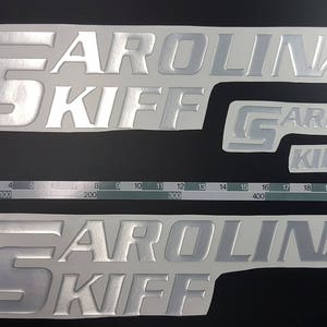 May include: Three silver-colored vinyl decals with the text "CAROLINA KIFF" in a raised, embossed font. The decals are on a white background.