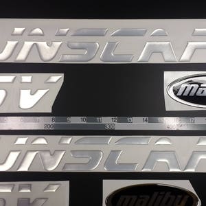 Puede incluir: Calcomanías plateadas SUNSCAPE y LSV, junto con emblemas ovalados Malibu. Las calcomanías están sobre un respaldo blanco con una regla en centímetros y pulgadas para la escala. El texto es en negrita y metálico.