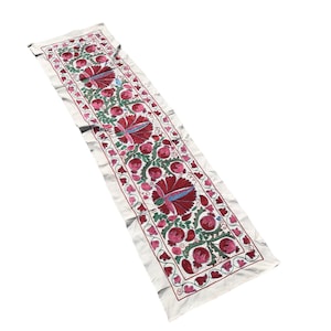 May include: White fabric table runner with a repeating pattern of embroidered red, pink, green, and blue flowers.