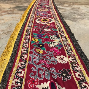 Pode incluir: Um longo corredor têxtil retangular vermelho com intrincados bordados florais em várias cores. As bordas são adornadas com bordas pretas e douradas e uma franja dourada. O corredor está colocado sobre uma superfície de concreto.