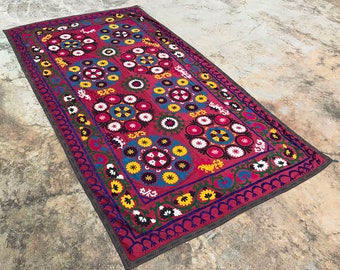 Puede incluir: Una alfombra grande, rectangular, roja bordada con un patrón floral. La alfombra tiene un borde marrón oscuro y está decorada con flores y círculos coloridos.
