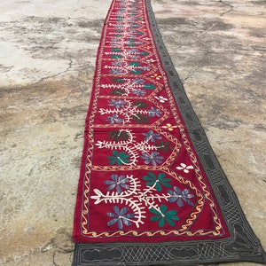 May include: A long, red fabric runner with intricate embroidery featuring floral designs in white, blue, and green. The runner has a black border with a geometric pattern.