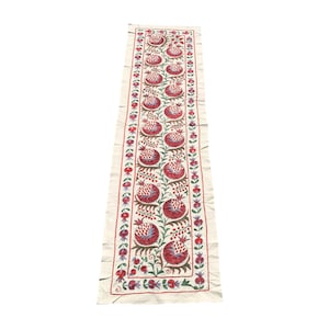 May include: A long white runner with a repeating pattern of red and green embroidered pomegranates.