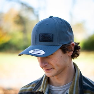 Puede incluir: Una persona que lleva una camisa a rayas grises y negras con una gorra de béisbol gris. La gorra tiene un parche de cuero negro con la palabra "Leup" en él. La gorra también tiene una pegatina blanca que dice "The Authentic Snapback".
