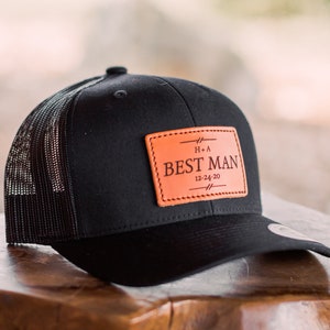May include: Black and brown trucker hat with a leather patch that says "Best Man" with the date "12-24-20" and the initials "H+A".