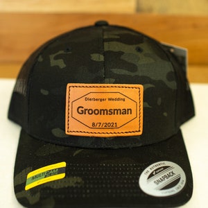 May include: A black and green camouflage trucker hat with a brown leather patch that says "Dierberger Wedding,  Groomsman, 8/7/2021".