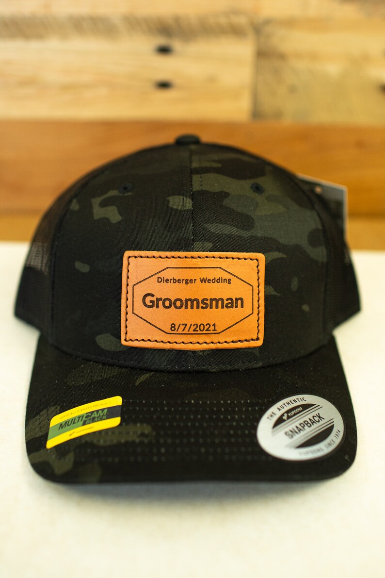 May include: A black and green camouflage trucker hat with a brown leather patch that says "Dierberger Wedding,  Groomsman, 8/7/2021".