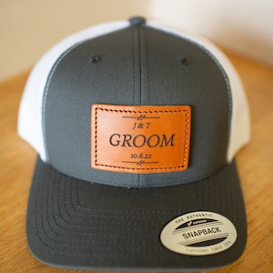 May include: A gray and white baseball cap with a brown leather patch that says "J&T GROOM 10.8.22". The cap has a white mesh back and a black "The Authentic Yupoong Snapback" label.