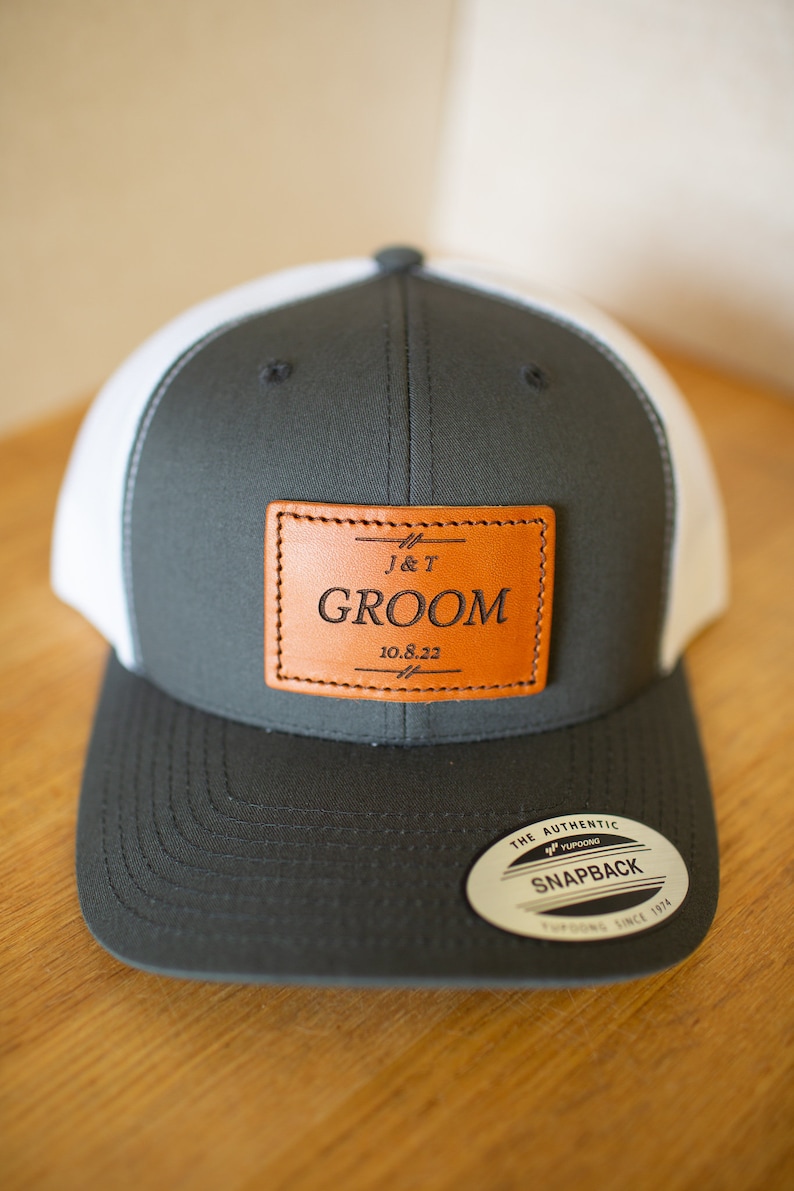 May include: A gray and white baseball cap with a brown leather patch that says "J&T GROOM 10.8.22". The cap has a white mesh back and a black "The Authentic Yupoong Snapback" label.
