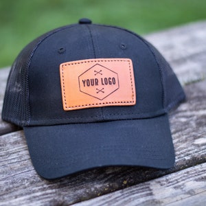 May include: Black trucker hat with a brown leather patch that says "YOUR LOGO" in a hexagon shape.