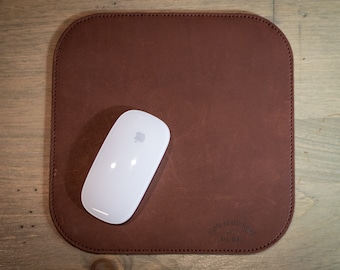 May include: A brown leather mouse pad with a stitched border and the text "Craft Wood Shop Est. 1982" in the bottom right corner. A white wireless mouse is on the pad.