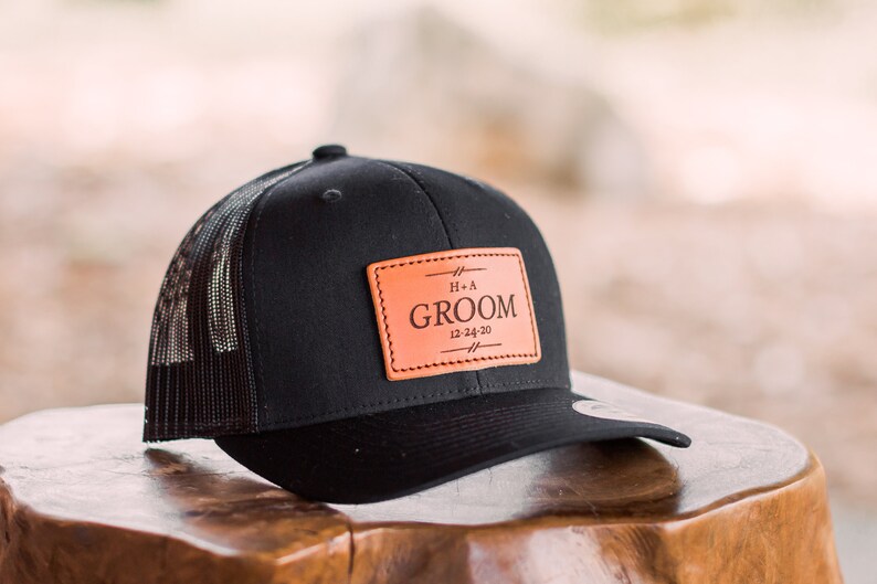 May include: Black trucker hat with a brown leather patch that says "Groom" with the date "12-24-20" and the initials "H+A" above the word "Groom".
