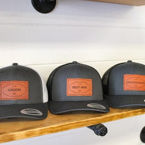 May include: Three gray and black trucker hats with brown leather patches. The patches have white text that reads "Groom", "Best Man", and "Groomsman".