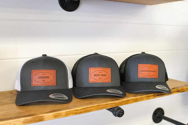 May include: Three gray and black trucker hats with brown leather patches. The patches have white text that reads "Groom", "Best Man", and "Groomsman".