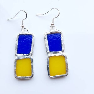 May include: A pair of handmade earrings featuring a blue and yellow stained glass design. Each earring has a rectangular shape with a blue top and a yellow bottom, connected by a silver-colored metal frame and a silver hook.