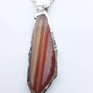 May include: A silver chain necklace with a pendant featuring a red, white, and brown agate stone set in a silver frame.