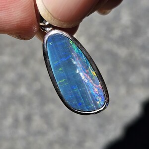 Peut inclure: Un pendentif en forme de goutte d'eau serti d'une opale bleu foncé aux reflets irisés verts, rouges et jaunes. La pierre est enchâssée dans une monture en métal argenté lisse et polie, dotée d'une bélière sur la partie supérieure.