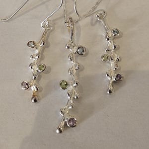 May include: A silver jewellery set consisting of a pendant on a chain and two matching drop earrings. Each piece features a textured, branch-like design adorned with small, round, multi-coloured gemstones in shades of light blue, green, and purple.