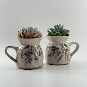 May include: Two small, speckled ceramic mugs used as planters, each featuring a hand-painted pattern of blue leaves and red berries. Each mug contains a green succulent plant, with the left succulent showing reddish tips on its leaves.