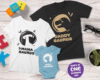 Op de afbeelding: Drie bijpassende T-shirts met een dinosaurus thema. De volwassen shirts zijn zwart en wit met de tekst "Daddy Saurus" en "Mama Saurus". Het baby shirt is lichtblauw met de tekst "Baby Saurus".