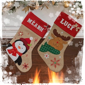 Puede incluir: Dos calcetines navideños de arpillera con puños rojos. El calcetín izquierdo presenta un pingüino con gorro y bufanda rojos, personalizado con el nombre MÉABH. El derecho muestra un reno con una bufanda de cuadros verdes, personalizado con el nombre LUCY.