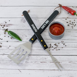May include: Two black handled grilling tools, a spatula and a fork, with the words "Patrick" and "Dad's BBQ" on the handles. The tools are crossed over each other on a white wooden surface. There is a small red chili pepper, a green jalapeno pepper, and a small container of red sauce on the surface.