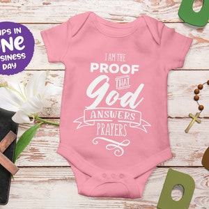 Könnte beinhalten: Rosaer Baby-Strampler mit weißem Text, der "Ich bin der Beweis, dass Gott Gebete erhört" lautet.