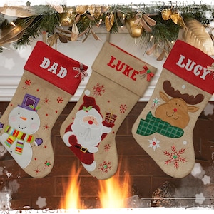 Puede incluir: Tres calcetines de Navidad personalizados de tela de arpillera con tapas de fieltro rojo. Los calcetines están decorados con copos de nieve bordados, un muñeco de nieve, Papá Noel y un reno. Los calcetines están colgados en una repisa de chimenea con una guirnalda de vegetación y luces.