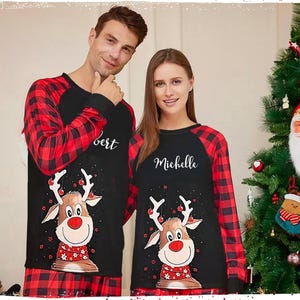 May include: A couple wearing matching black and red Christmas pyjamas with reindeer designs. The man's pyjamas say "Bert" and the woman's pyjamas say "Michelle".