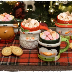 Könnte beinhalten: Vier festliche Tassen gefüllt mit Marshmallows, jede als andere Weihnachtsfigur gestaltet: ein Rentier, der Weihnachtsmann, ein Schneemann und ein Pinguin. Die Tassen stehen auf einer rot-schwarz karierten Oberfläche, mit Keksen in der Nähe.
