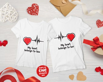 Puede incluir: Dos camisetas blancas con un corazón rojo y una línea de ECG negra. El texto en las camisetas dice "Mi corazón le pertenece a él" y "Mi corazón le pertenece a ella".