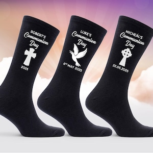 Può includere: Tre paia di calzini neri con disegni bianchi. I calzini presentano una croce, una colomba e una croce celtica. Ogni calzino ha un nome e una data stampati. Il testo sui calzini recita "Robert's Communion Day 2025", "Luke's Communion Day 6th May 2025" e "Micheal's Communion Day 22.05.2025".