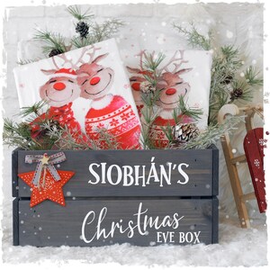 Könnte beinhalten: Eine graue Holzkiste für Weihnachten mit dem weißen Text "SIOBHÁN'S Christmas EVE BOX". Die Kiste enthält weihnachtliche Servietten mit Rentier-Designs, Tannennadeln und einen roten Stern.