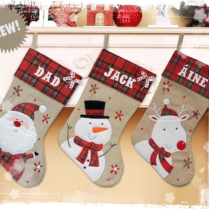Peut inclure: Trois chaussettes de Noël personnalisées avec une bordure en tartan rouge et vert. Les chaussettes présentent des images brodées de Père Noël, d'un bonhomme de neige et d'un renne. Les chaussettes sont étiquetées "Papa", "Jack" et "Aine".