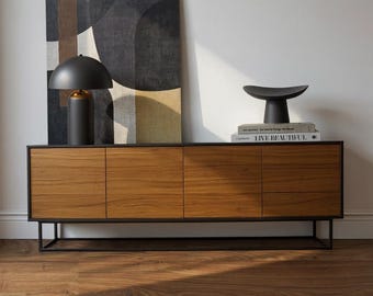 May include: A contemporary living room setting with a wooden cabinet with four doors, a black metal frame, and a black lamp with a gold base. A black sculpture and books are on top of the cabinet. An abstract painting hangs on the wall.
