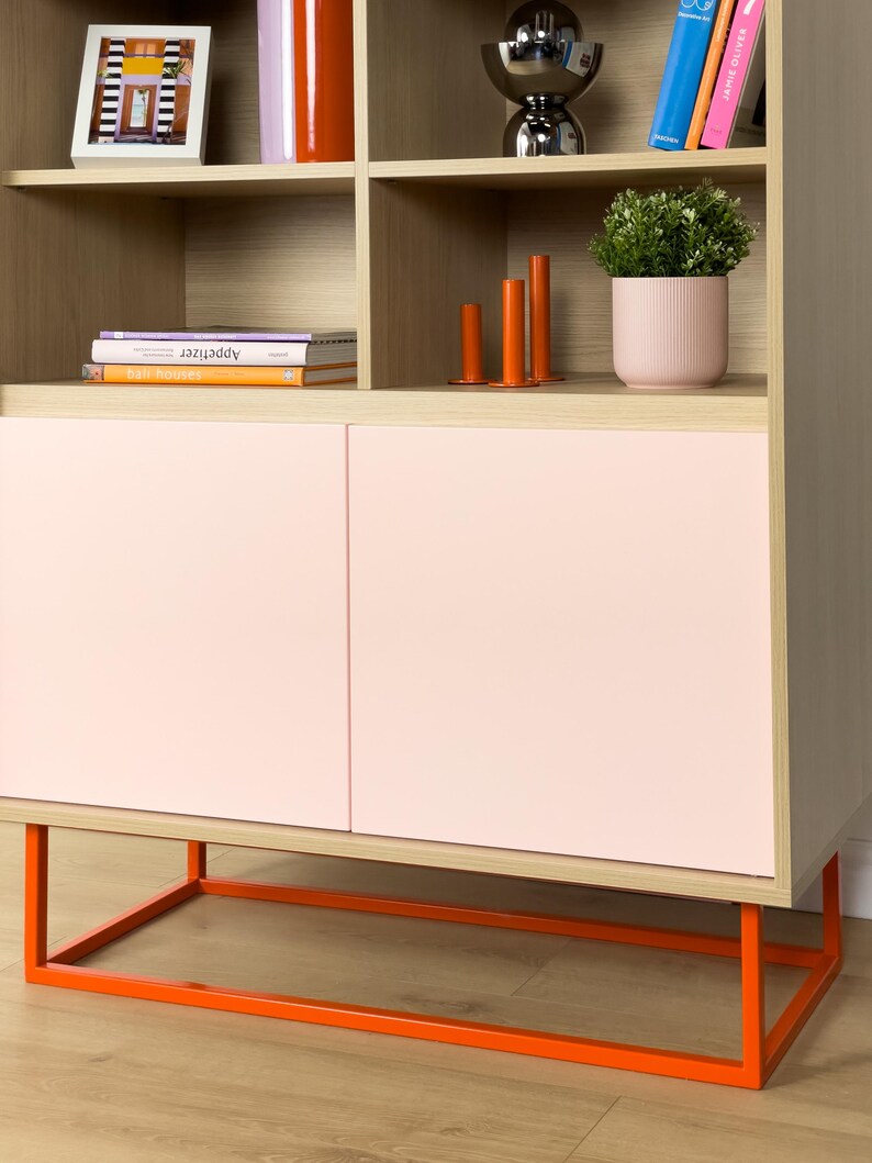 May include: A modern wooden cabinet with light pink doors and an orange metal base. The cabinet has open shelves with decorative items, including books, vases, and a potted plant. The overall design is minimalist and stylish.
