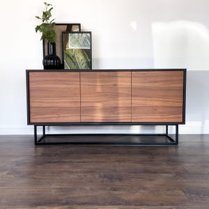 Könnte beinhalten: Ein moderner Holzschrank mit drei Türen, mit dunkler Holzmaserung und einem schwarzen Metallrahmen. Der Schrank ist mit einer schwarzen Vase und gerahmten Kunstwerken dekoriert. Er steht auf einem dunklen Holzboden.
