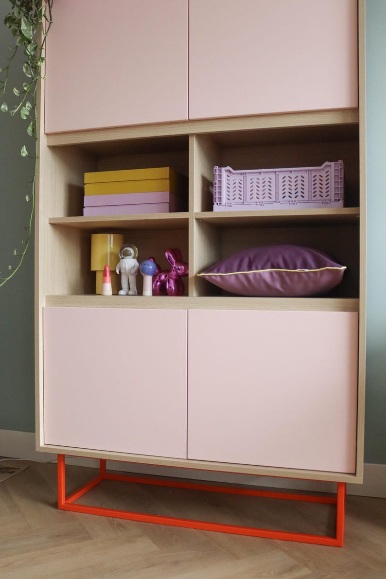 May include: A modern storage cabinet with a light pink finish and orange metal base. The cabinet features open shelves with decorative items like boxes, a crate, and figurines. A purple pillow with yellow trim rests on a shelf.