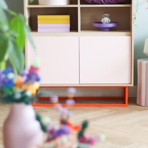 May include: A light pink cabinet with open shelves and closed doors. The cabinet has a natural wood frame and orange base. Shelves display a disco ball, purple pillow, and stacked boxes. A vase of flowers is in the foreground.