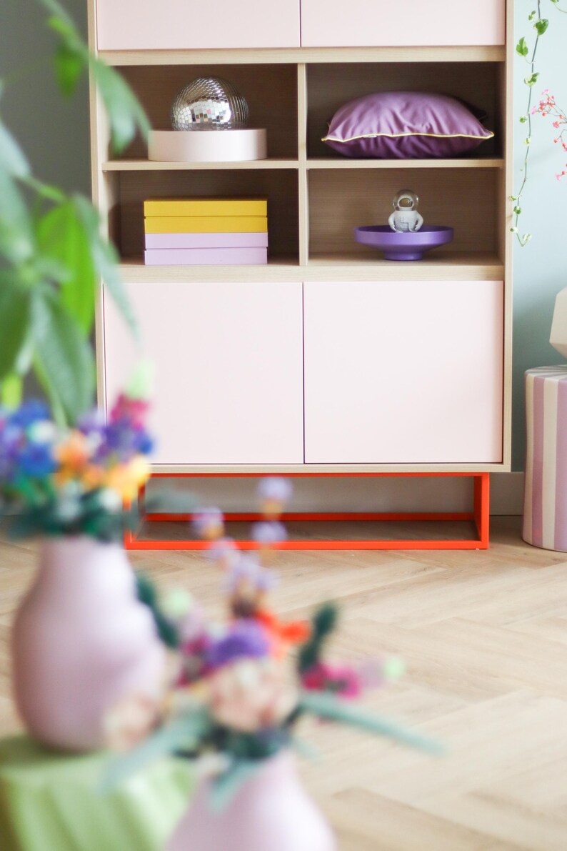 May include: A light pink cabinet with open shelves and closed doors. The cabinet has a natural wood frame and orange base. Shelves display a disco ball, purple pillow, and stacked boxes. A vase of flowers is in the foreground.