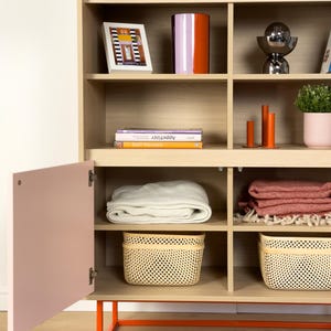 May include: A light wood cabinet with open shelves and a pink door. The shelves display decorative items like a framed picture, a vase, books, and baskets. The cabinet has an orange metal base and is filled with home decor.
