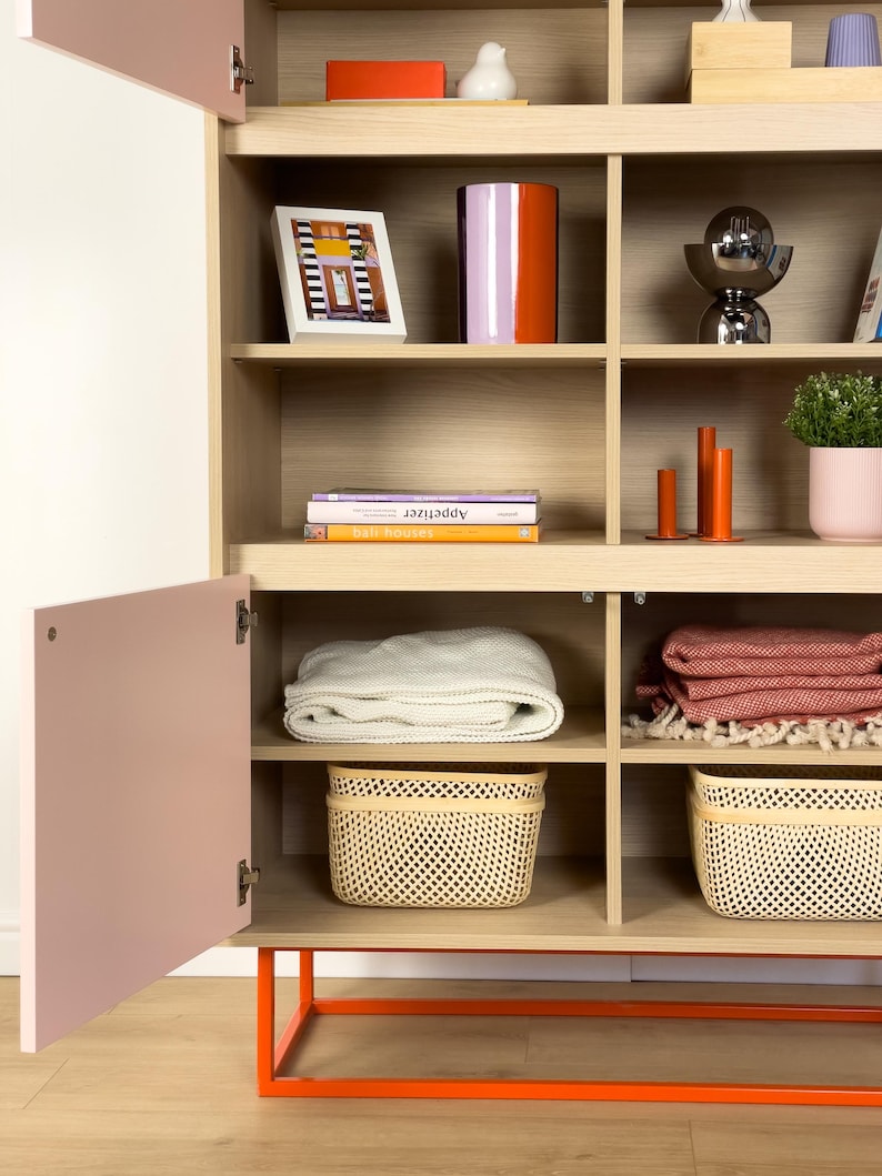May include: A light wood cabinet with open shelves and a pink door. The shelves display decorative items like a framed picture, a vase, books, and baskets. The cabinet has an orange metal base and is filled with home decor.