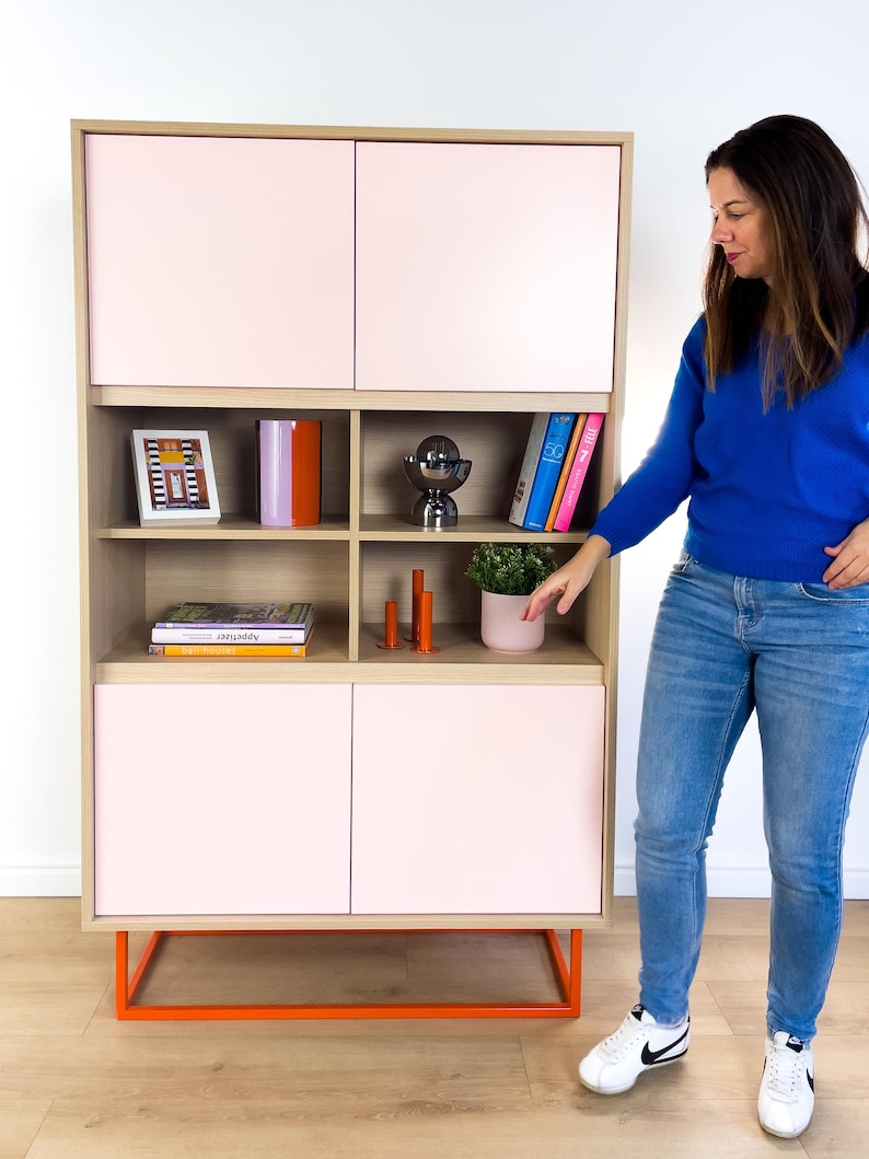 May include: A modern wooden cabinet with light pink doors and open shelving. The cabinet has an orange metal base and is filled with books, decorative items, and a small potted plant. The cabinet is against a white wall.