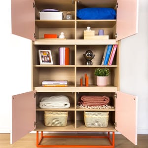 May include: A modern wooden cabinet with light pink doors and an orange base. The cabinet is filled with various items, including a blue pillow, books, decorative objects, blankets, and woven baskets. The cabinet is open, revealing the contents.