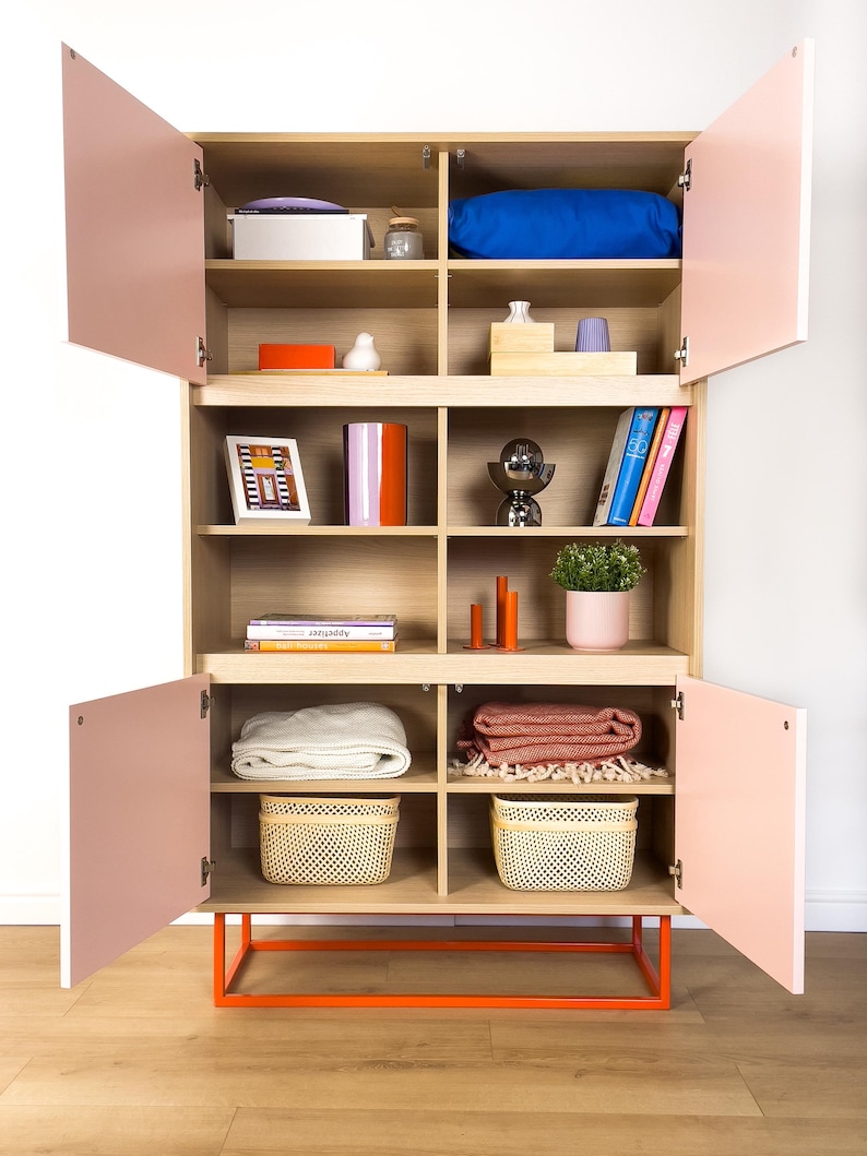 May include: A modern wooden cabinet with light pink doors and an orange base. The cabinet is filled with various items, including a blue pillow, books, decorative objects, blankets, and woven baskets. The cabinet is open, revealing the contents.